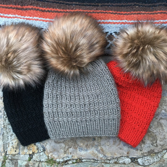 Three beanies colored black, gray, and red all with brown poms against a stone background and red falsa blanket.