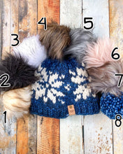 Load image into Gallery viewer, Snowflake Beanie in navy showing all pom options: cream, black, white, brown, gray/black, pink, gray, matching yarn. It is shown in a flat lay against a wooden background.

