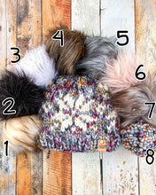 Load image into Gallery viewer, Snowflake Beanie in gray showing all pom options: cream, black, white, brown, gray/black, pink, gray, matching yarn. It is shown in a flat lay against a wooden background.
