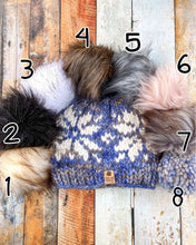 Load image into Gallery viewer, Snowflake Beanie in light blue showing all pom options: cream, black, white, brown, gray/black, pink, gray, matching yarn. It is shown in a flat lay against a wooden background.
