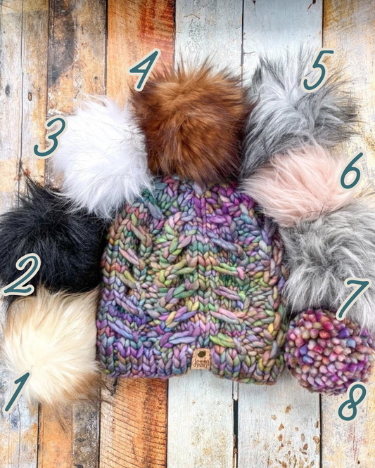 Fossil Beanie in rainbow shown in a flat lay with all 8 pom options.