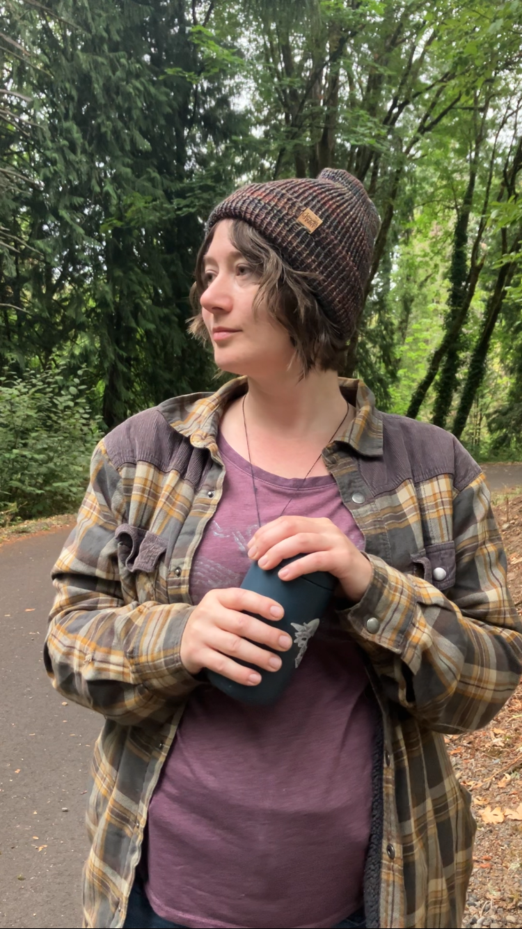 The Hadrosaur Hat is a lightweight, double layered slouchy beanie with folded brim. It is shown here in dark rainbow on a model on a hiking trail.