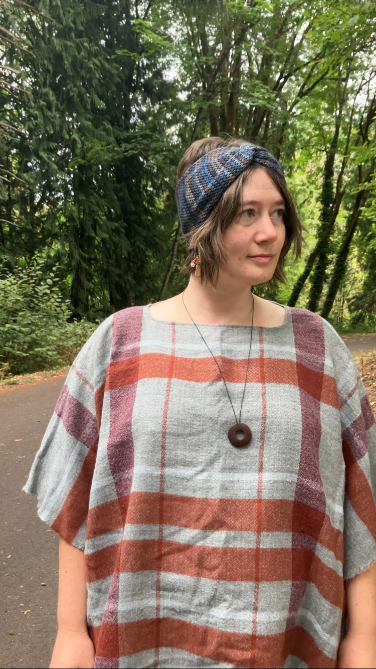 Knitted headband with knotted front shown on a model in the woods. The headband is a blend of light blues, grays, and browns.