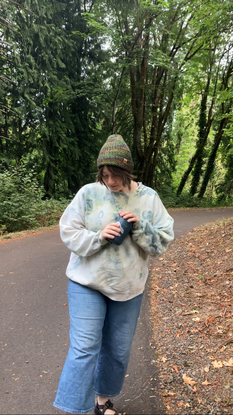 The Hadrosaur Hat is a lightweight, double layered slouchy beanie with folded brim. It is shown here in light rainbow on a model on a hiking trail.