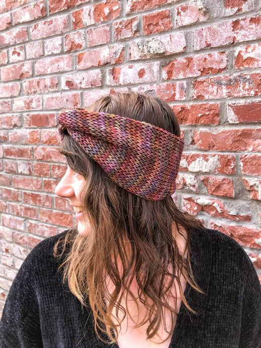 Knitted headband with knotted front shown on a model.  The headband is brown, purple, and green, and the model's head is turned to the left.