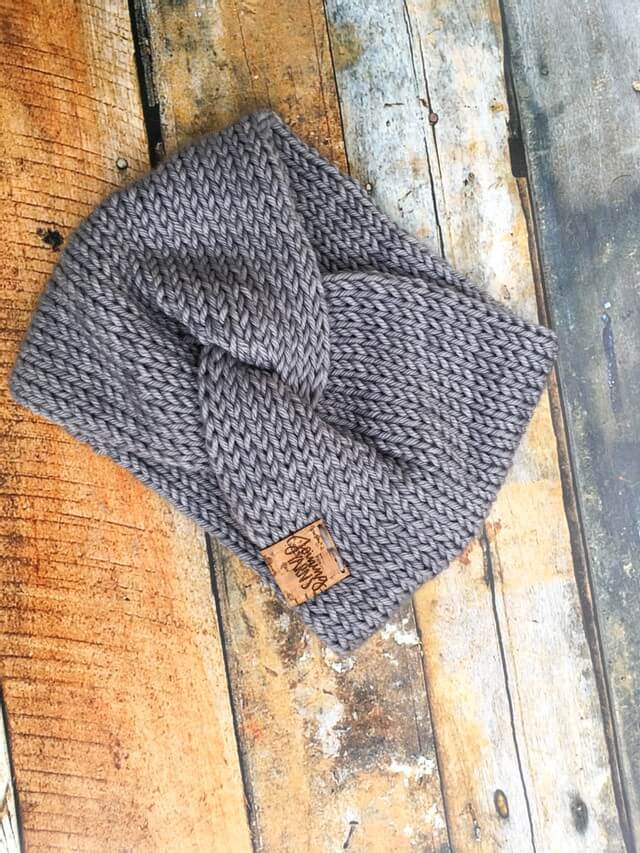 A knit headband with knot in the front shown against a wooden background. The headband is a light gray.