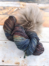 Load image into Gallery viewer, Skein of dark rainbow yarn with gray pom.
