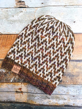 Load image into Gallery viewer, The Find Your Way Beanie has a chevron stripe pattern in two colors.  It is shown here in a flat lay in white and brown.
