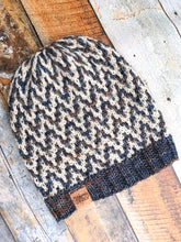 Load image into Gallery viewer, The Find Your Way Beanie has a chevron stripe pattern in two colors.  It is shown here in a flat lay in white and blue/brown.

