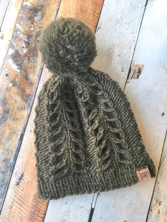 An olive colored fossil beanie with yarn pom in a flat lay against a wooden background.