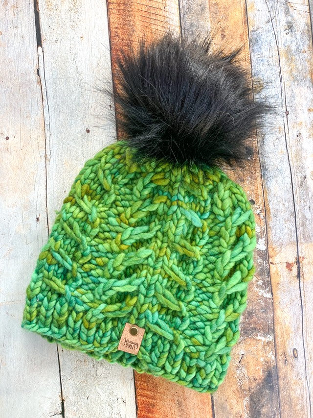 A chunky fossil beanie in light green with black pom in a flat lay against a wood background.