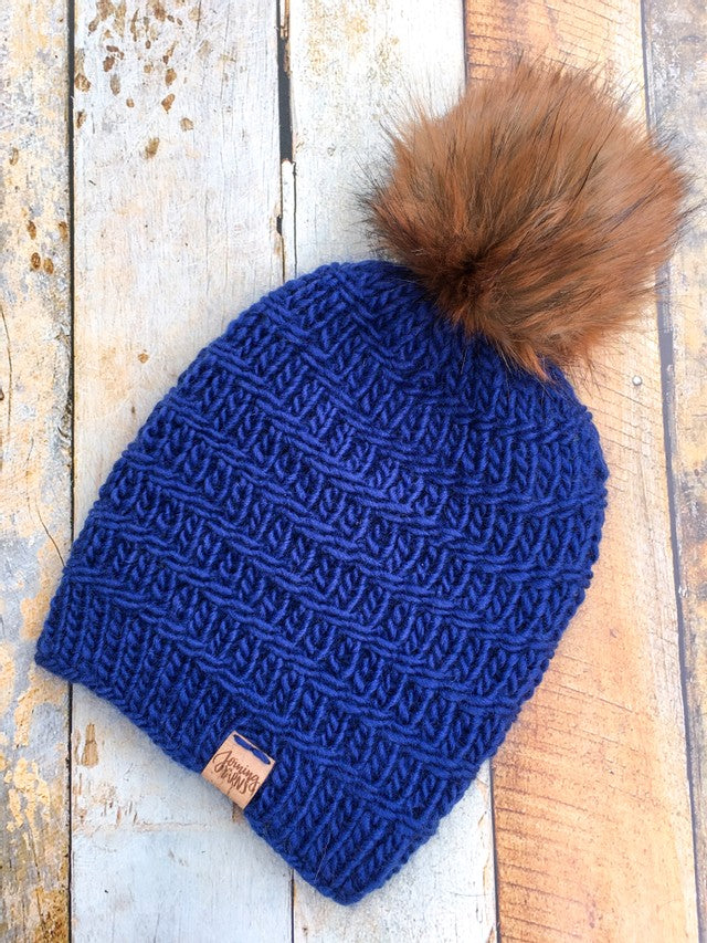 A navy blue Messenger Beanie with brown pom in a flt lay against a wood background.