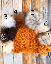 Load image into Gallery viewer, Fossil Beanie in orange showing all pom options:  cream, black, white, brown, gray/black, pink, gray, matching yarn.  It is shown in a flat lay against a wooden background.
