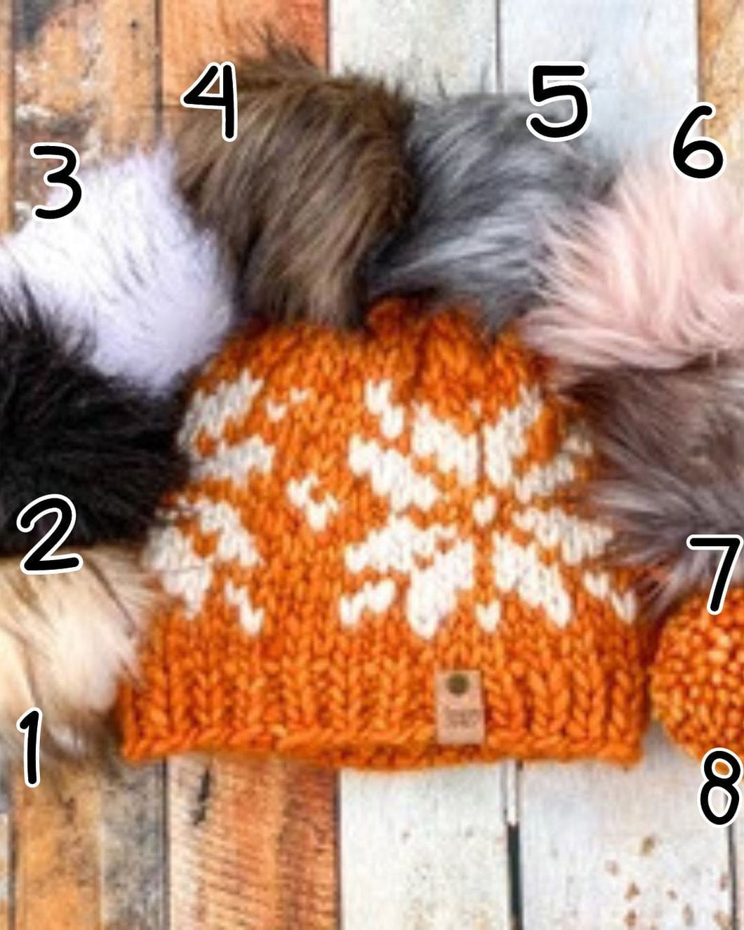 Snowflake Beanie in orange showing all pom options: cream, black, white, brown, gray/black, pink, gray, matching yarn. It is shown in a flat lay against a wooden background.