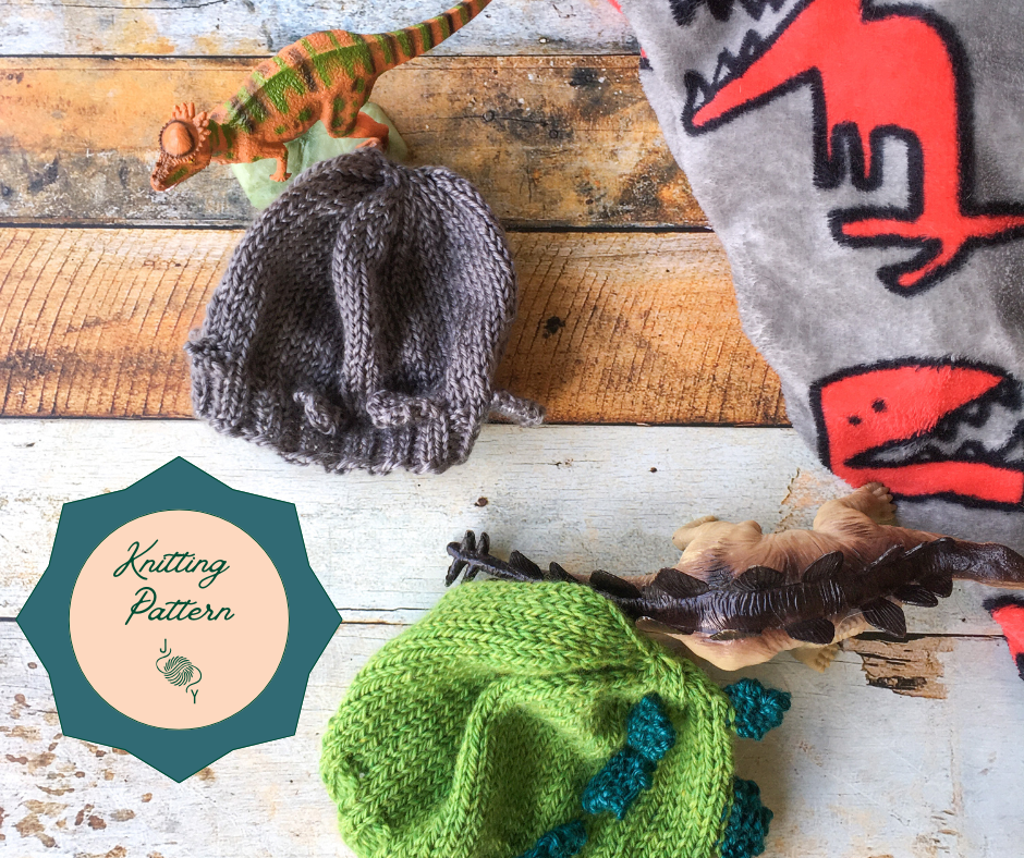 Two knitted baby hats lay against a fleece blanket with dinosaurs on it that is laying on a wooden background.  The green hat is styled after a stegosaurus and the gray hat after a pachycephalosaurus.  A circle with the words "knitting pattern" is next to them.