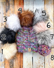 Load image into Gallery viewer, Witch Hazel Beanie in rainbow showing all pom options:  cream, black, white, brown, gray/black, pink, gray, matching yarn.  It is shown in a flat lay against a wooden background.
