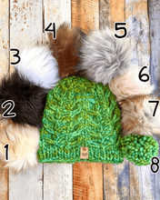 Load image into Gallery viewer, Fossil Beanie in green showing all pom options:  cream, black, white, brown, gray/black, pink, gray, matching yarn.  It is shown in a flat lay against a wooden background.
