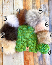 Load image into Gallery viewer, Witch Hazel Beanie in green showing all pom options:  cream, black, white, brown, gray/black, pink, gray, matching yarn.  It is shown in a flat lay against a wooden background.
