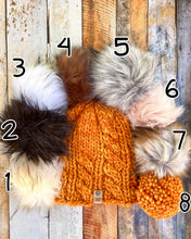 Load image into Gallery viewer, Witch Hazel Beanie in orange showing all pom options:  cream, black, white, brown, gray/black, pink, gray, matching yarn.  It is shown in a flat lay against a wooden background.
