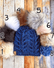 Load image into Gallery viewer, Fossil Beanie in dark blue showing all pom options:  cream, black, white, brown, gray/black, pink, gray, matching yarn.  It is shown in a flat lay against a wooden background.
