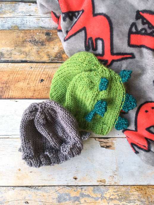 Two knitted baby hats lay against a fleece blanket with dinosaurs on it that is laying on a wooden background.  The green hat is styled after a stegosaurus and the gray hat after a pachycephalosaurus.