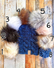 Load image into Gallery viewer, Witch Hazel Beanie in dark blue showing all pom options:  cream, black, white, brown, gray/black, pink, gray, matching yarn.  It is shown in a flat lay against a wooden background.
