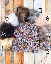 Load image into Gallery viewer, Fossil Beanie in gray showing all pom options: cream, black, white, brown, gray/black, pink, gray, matching yarn. It is shown in a flat lay against a wooden background.

