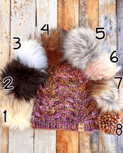 Load image into Gallery viewer, Fossil Beanie in brown showing all pom options:  cream, black, white, brown, gray/black, pink, gray, matching yarn.  It is shown in a flat lay against a wooden background.
