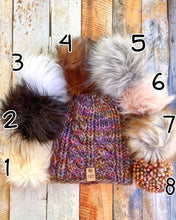 Load image into Gallery viewer, Witch Hazel Beanie in brown showing all pom options:  cream, black, white, brown, gray/black, pink, gray, matching yarn.  It is shown in a flat lay against a wooden background.
