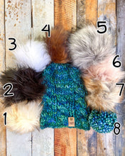 Load image into Gallery viewer, Witch Hazel Beanie in teal showing all pom options:  cream, black, white, brown, gray/black, pink, gray, matching yarn.  It is shown in a flat lay against a wooden background.
