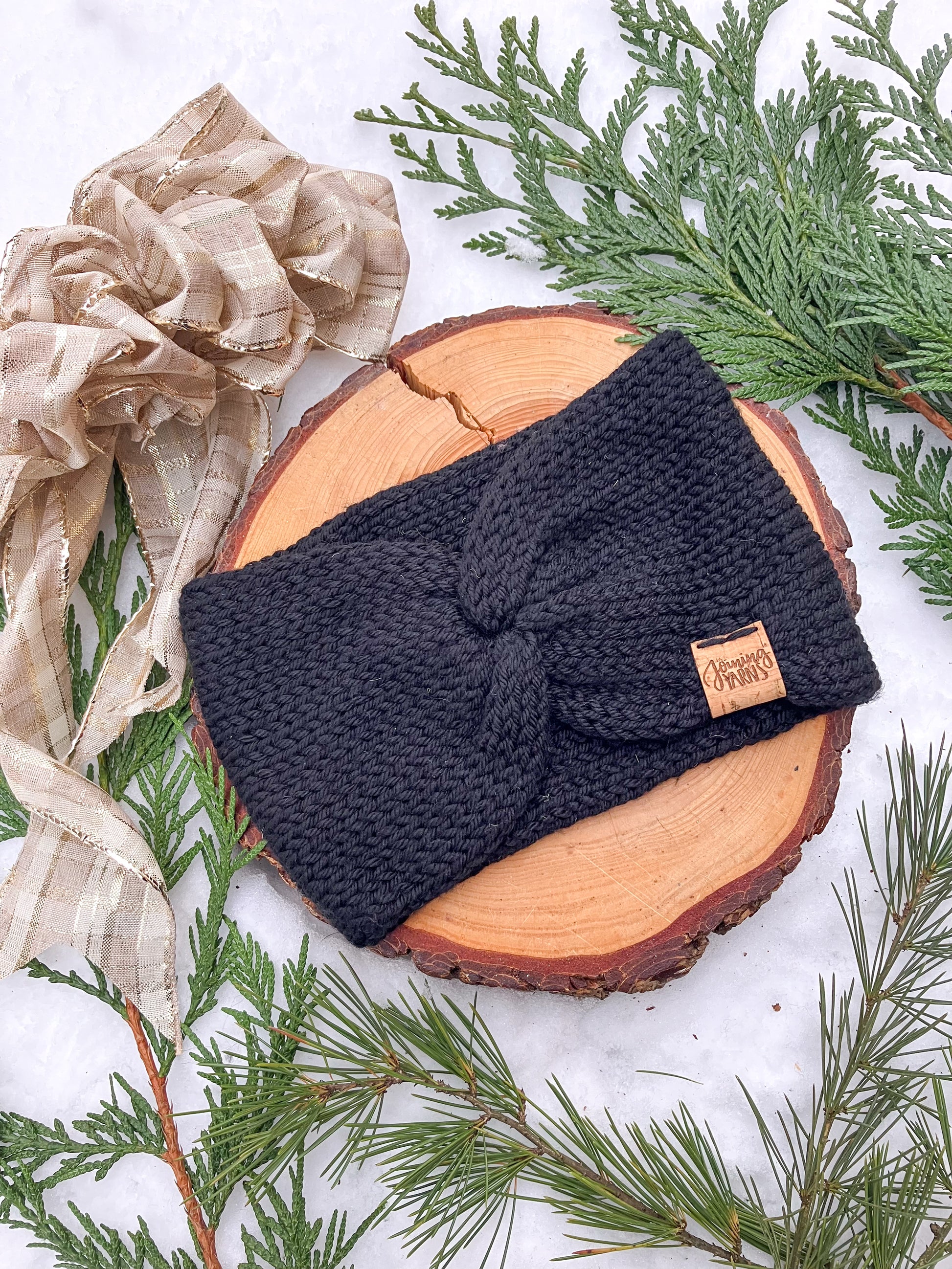Flat lay of an Ankylosaur Headwrap in black against a wood circle in the snow surrounded by evergreen branches.