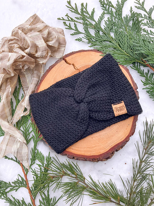Flat lay of an Ankylosaur Headwrap in black against a wood circle in the snow surrounded by evergreen branches.
