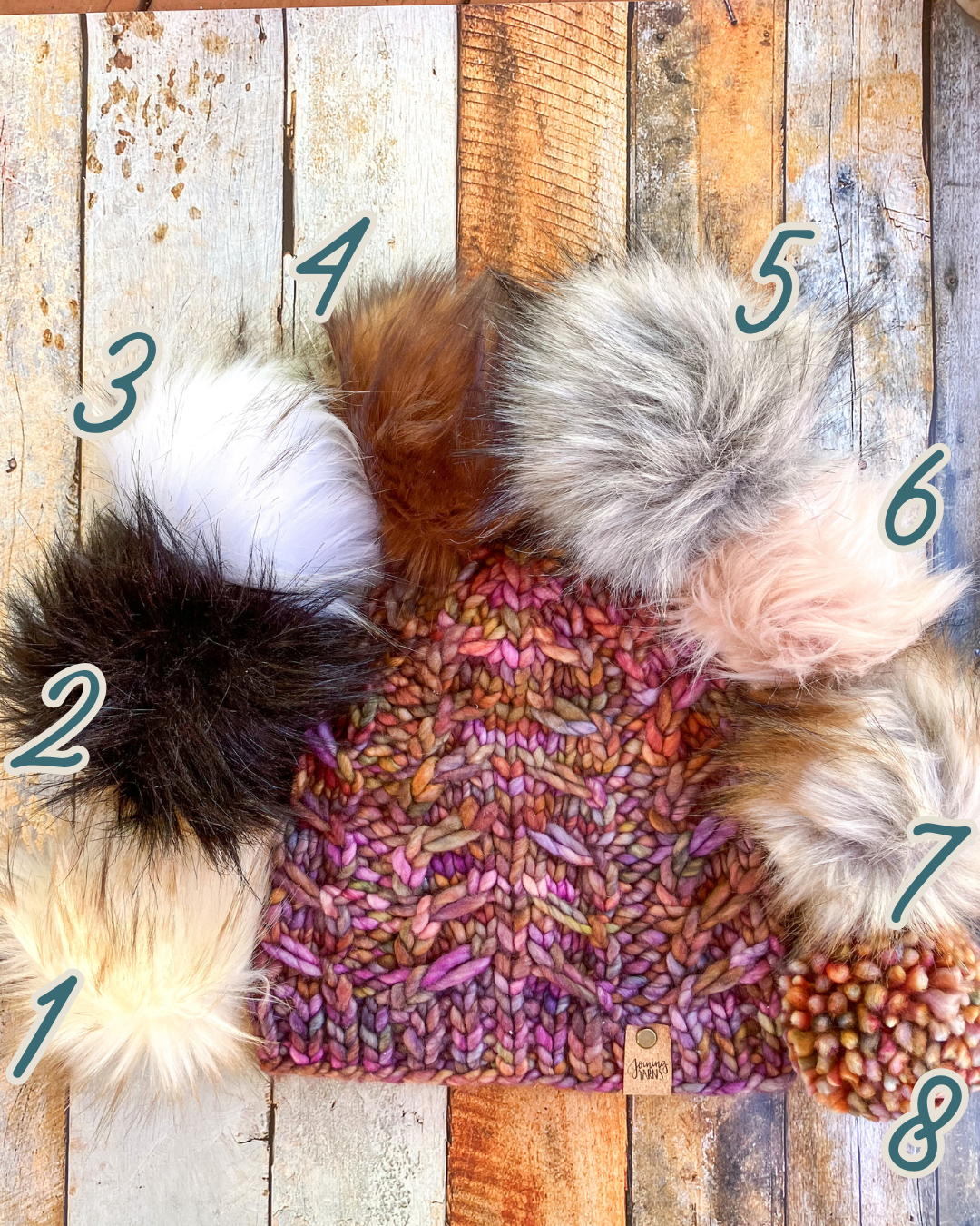 Fossil Beanie in brown shown in a flat lay with all 8 pom options.