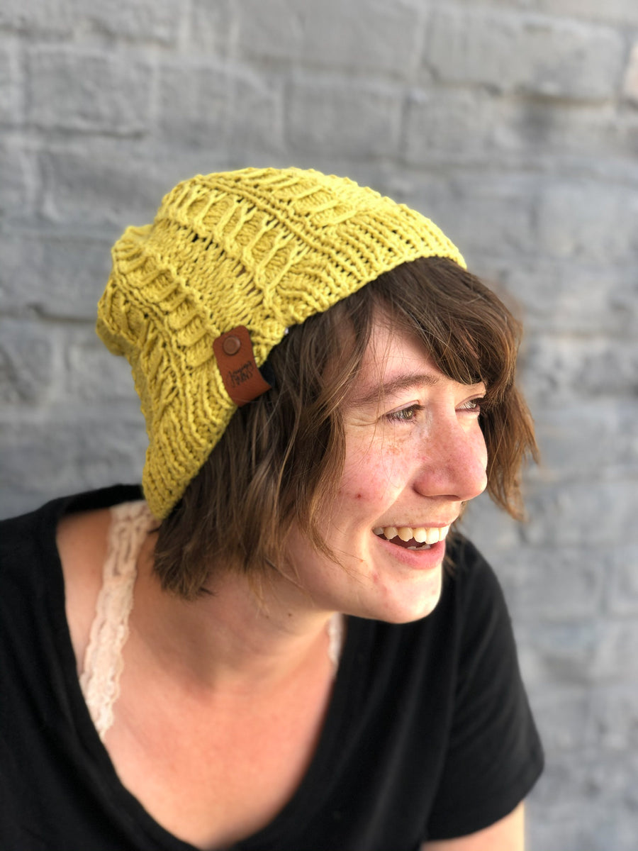 Photo of business owner in yellow slouchy beanie and black shirt against blue brick background.