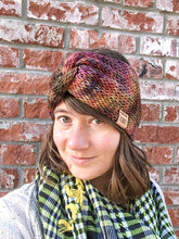 Load image into Gallery viewer, Knitted headband with knotted front shown on a model against a brick background.  The headband in a blend of pinks, golds, and purples.
