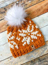 Load image into Gallery viewer, Snowflake Beanie in orange with white snowflake and white pom.  Shown here in a flat lay against a wooden background.
