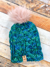 Load image into Gallery viewer, Witch Hazel Beanie in teal with pink pom. It is shown here in a flat lay against a wooden background.
