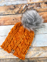 Load image into Gallery viewer, Witch Hazel Beanie in orange with gray pom. It is shown here in a flat lay against a wooden background.
