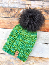 Load image into Gallery viewer, Witch Hazel Beanie in green with black pom. It is shown here in a flat lay against a wooden background.
