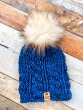 Load image into Gallery viewer, Witch Hazel Beanie in dark blue with cream pom. It is shown here in a flat lay against a wooden background.
