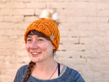 Load image into Gallery viewer, Witch Hazel Beanie in orange with cream pom. It is shown here on a model.
