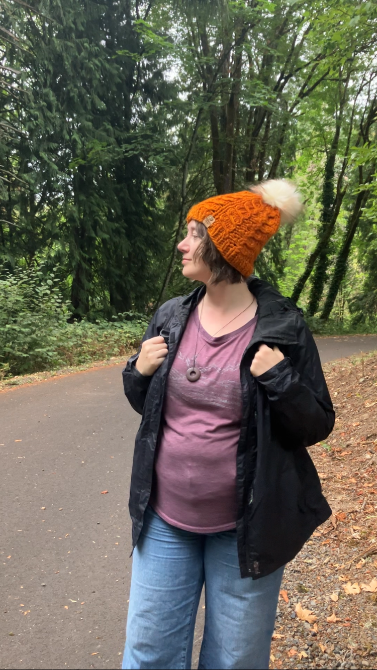 Fossil Beanie in orange with cream pom.  It is shown here on a model on a hiking trail.