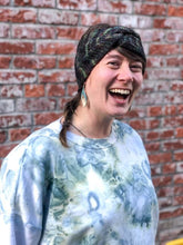 Load image into Gallery viewer, Knitted headband with knotted front shown on a model against a brick background.  The headband in a blend of dark rainbow colors, mostly green.
