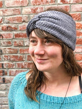 Load image into Gallery viewer, Knitted headband with knotted front shown on a model against a brick background.  The headband is light gray.

