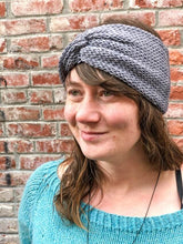 Load image into Gallery viewer, Knitted headband with knotted front shown on a model against a brick background.  The headband is light gray.
