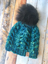 Load image into Gallery viewer, Fossil Beanie in green/blue with black pom.  It is shown here in a flat lay against a wooden background.

