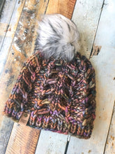 Load image into Gallery viewer, Fossil Beanie in brown with white pom with black streaks.  It is shown here in a flat lay against a wooden background.
