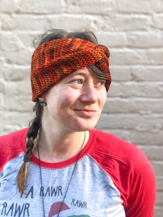 Knitted headband with knotted front shown on a model against a white brick background.  The headband is a blend of reds and oranges.