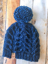 Load image into Gallery viewer, Fossil Beanie in dark blue with matching yarn pom.  It is shown here in a flat lay against a wooden background.
