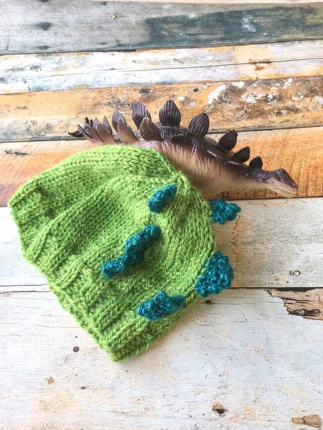 A green knitted baby hat styled after a stegosaurus lays against a wooden background with a toy stegosaurus next to it.